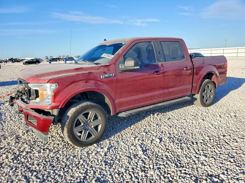 A white 2022 Ford F-150 XLT SuperCrew pickup truck with crumpled hood and front bumper damage at an auto body shop with chain-link fence